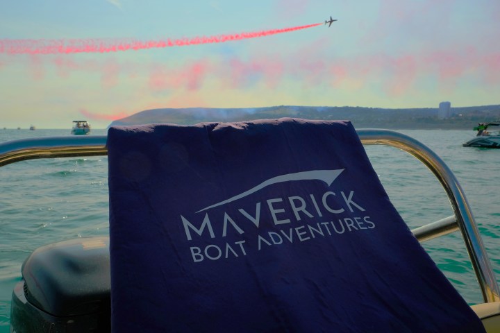 Boat on water with 'Maverick Boat Adventures' towel, plane with pink smoke in sky.