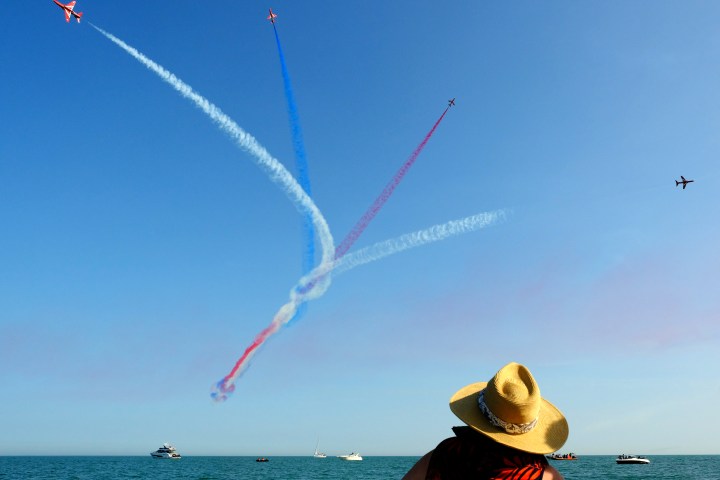 Person wearing a straw hat watches airplanes creating colorful trails across a clear sky over a calm sea.