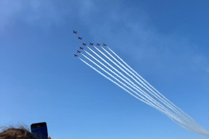 Person photographing jet formation flying over water with white vapor trails.