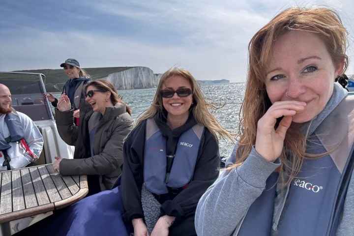 Group on a boat wearing life jackets, with cliffs and sea in the background.