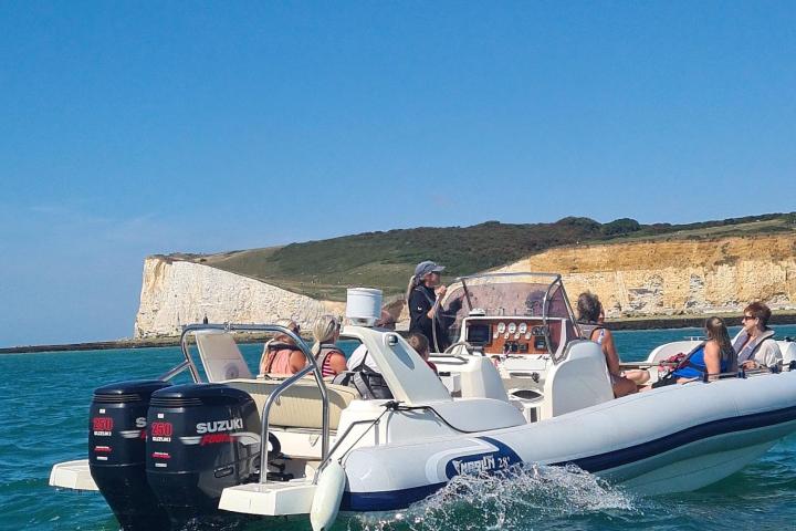 People on a boat with twin engines cruising near a rocky coastline under a clear blue sky.