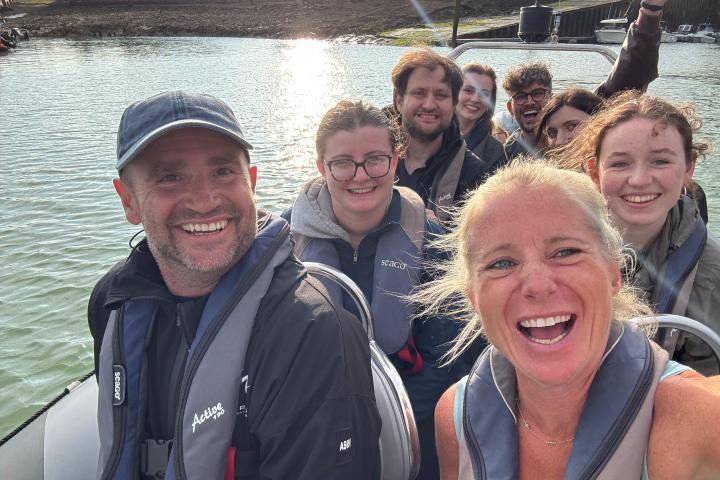 Group of smiling people on a boat by the water, wearing life jackets.