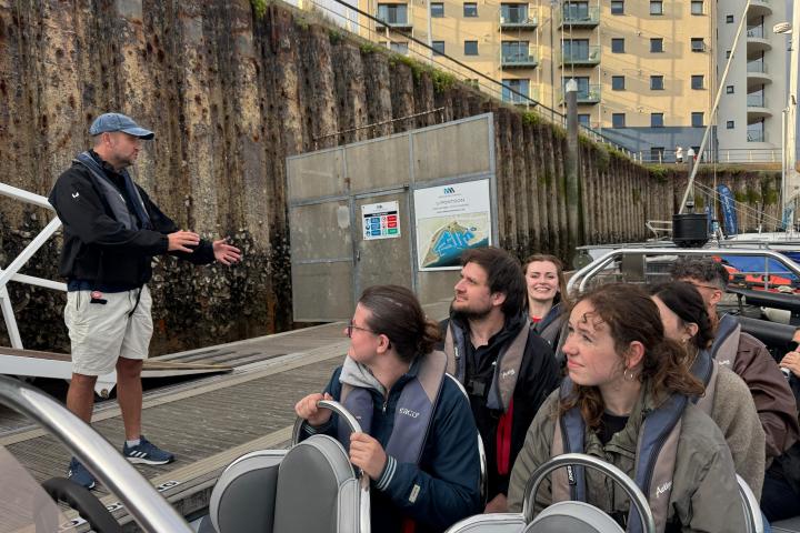 Group of people in a boat listening to a man speaking on a dock.