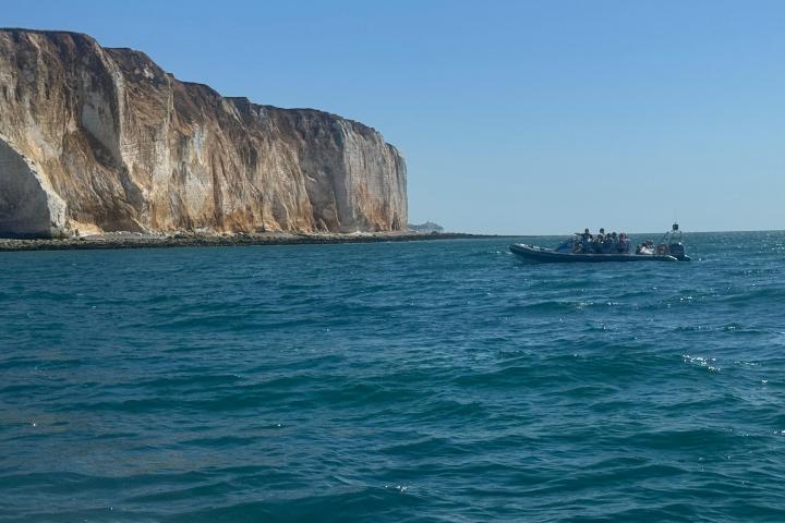 Speedboat with passengers near rugged cliffs over calm blue sea under clear sky.