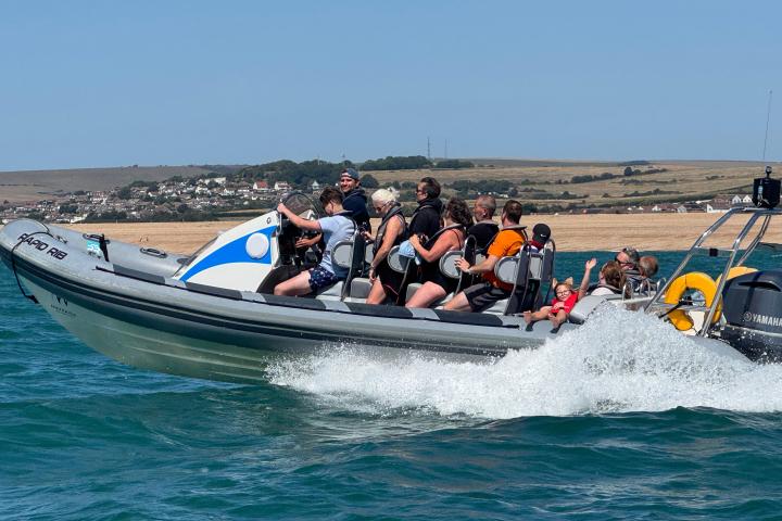 People in life jackets on a speedboat in blue waters near a coastal town.