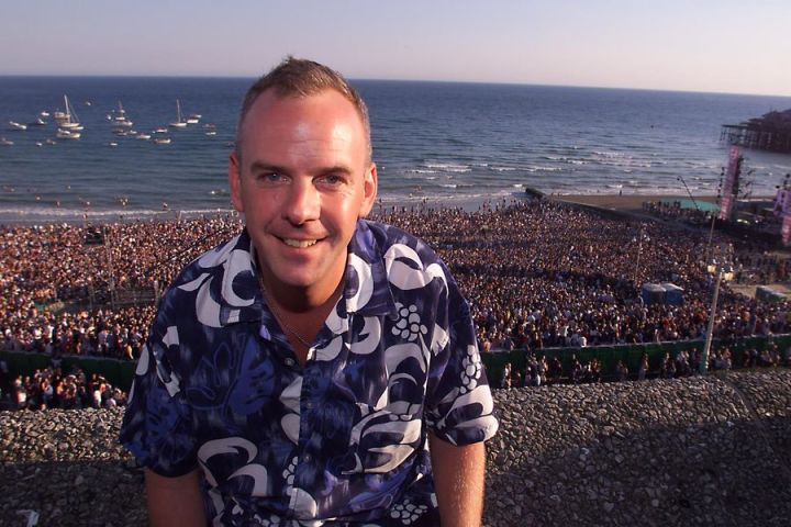Man smiling with a large beach crowd and ocean backdrop at sunset.